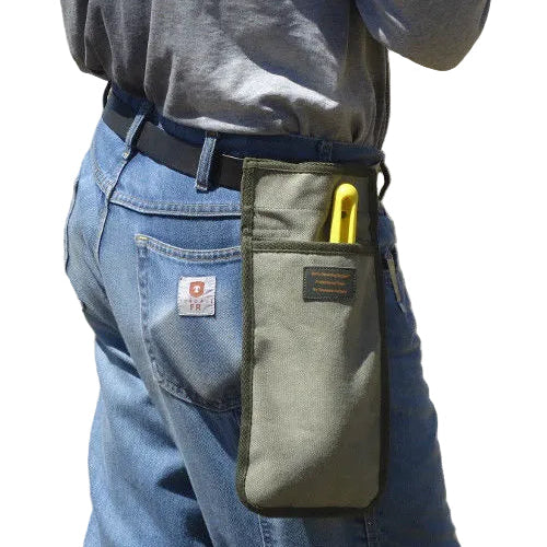 A close-up of a person outdoors wearing a gray canvas tool pouch on their waist belt, tucked into blue jeans. The pouch has a black trim and a yellow-handled tool inserted into it.